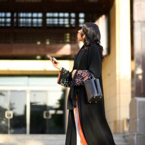 Side view shot of Saudi woman in colourful floral patterned abaya using smartphone near sunlit campus building