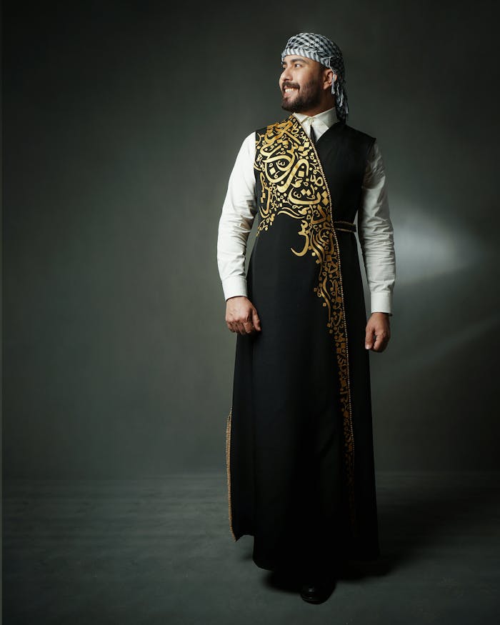 Studio portrait of a happy man wearing a thobe and kaffiyeh, standing against a dark background.