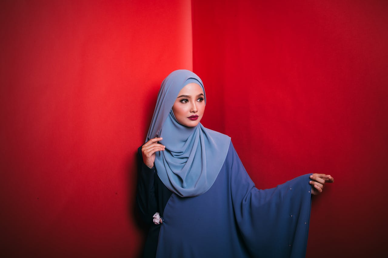 Pensive young ethnic female with red lips in traditional abaya and hijab standing in studio and looking away