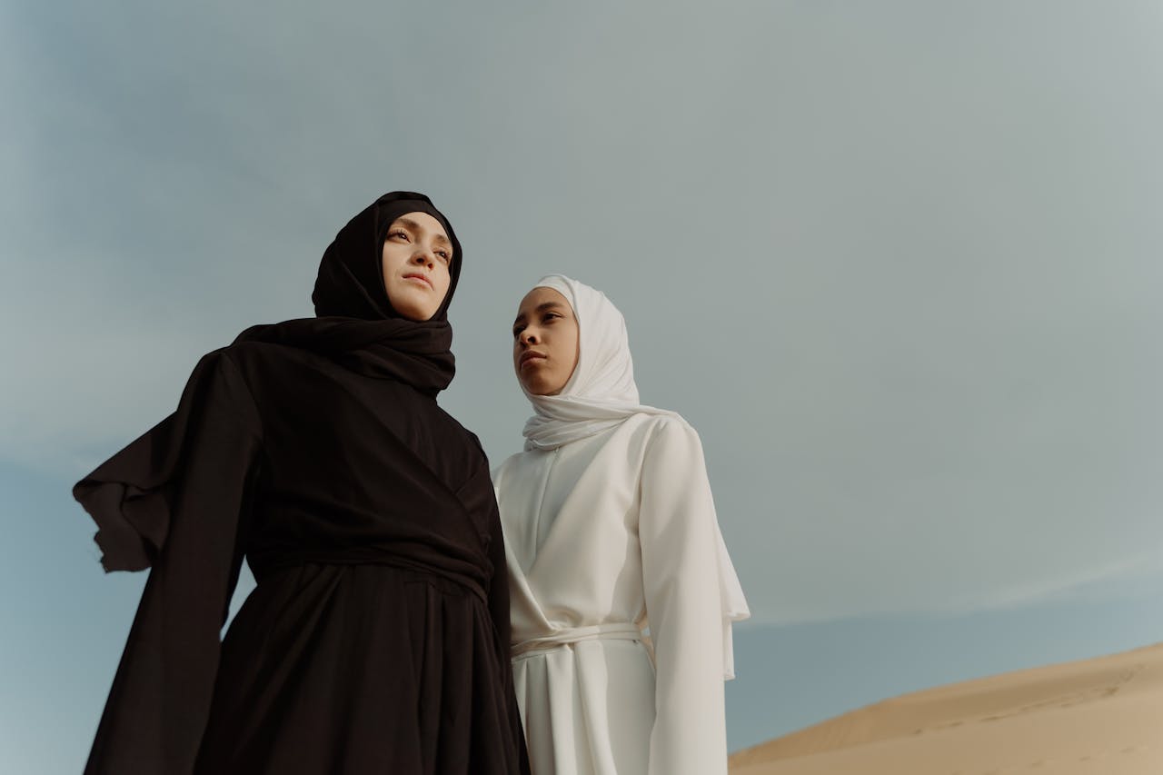Two women wearing elegant black and white hijabs stand confidently in a desert landscape, showcasing traditional attire.